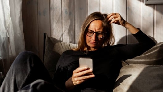 Woman with long hair and glasses sitting indoors looking down at a mobile phone